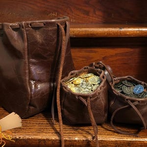 Pode incluir: Três bolsas de couro castanho escuro com cordão de tamanhos variados. As duas bolsas menores estão cheias de moedas metálicas em tons de ouro e prata. Uma ilustração gráfica de uma pena num tinteiro e um pergaminho enrolado é visível.