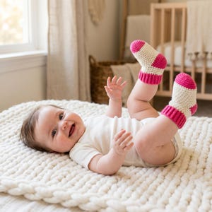 May include: A pair of cream and hot pink crocheted baby booties featuring a ribbed cuff and a rounded toe design. The booties are crafted with a thick yarn texture, showcasing a two-tone color block pattern with a solid cream base and vibrant pink accents on the toe, heel, and cuff.