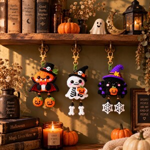 Puede incluir: Tres llaveros de ganchillo hechos a mano colgados de ganchos. A la izquierda, una calabaza vampiro con sombrero de copa negro, en el centro, un esqueleto con sombrero de bruja negro sosteniendo una calabaza, y a la derecha, una araña negra con sombrero morado.