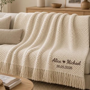 Könnte beinhalten: Eine cremefarbene Strickdecke mit einer strukturierten Noppenmuster-Optik und langen Fransen am unteren Rand. Die Decke ist mit dunkelbraunem Sticktext versehen, der Alice, ein Herzsymbol, Michael und 18.05.2026 zeigt.