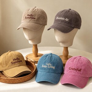 May include: Five baseball caps in various colours including tan, dark grey, mustard yellow, blue, and pink. Each cap features embroidered text: Hello Sunshine, Mountain Air, Coffee Easy Living, or Grateful. The caps have a washed, textured finish.