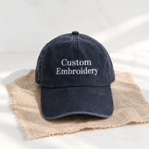 Può includere: Un cappellino da baseball grigio antracite scuro con una texture lavata e usurata. Il pannello frontale presenta il testo ricamato in bianco "Custom Embroidery" in un carattere serif pulito. Il cappellino ha una visiera curva e sei pannelli.
