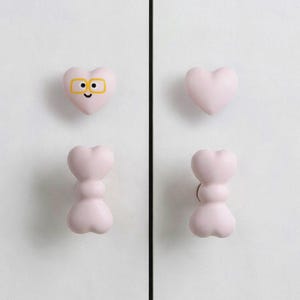 May include: Four light pink cabinet knobs mounted on white doors. Two knobs are heart-shaped, with one featuring a yellow-rimmed glasses graphic and a small black smile. The other two knobs have a stacked, double-lobed, bone-like silhouette.