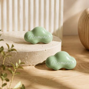 May include: Two smooth, sage green, cloud-shaped objects made of a solid material. Each piece features the name Ramona engraved in a white, cursive script font across the centre. The items have a glossy finish and rounded, organic edges.