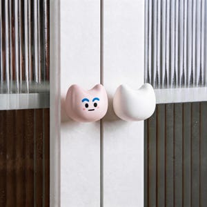 May include: Two cat-shaped cabinet knobs mounted on white cabinet doors. One knob is light pink with blue eyebrows, black eyes, and a small black mouth. The other knob is solid white. Both knobs have rounded ears and a smooth, matte finish.