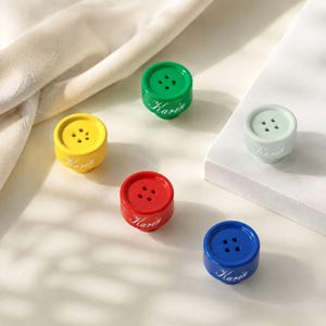 Puede incluir: Cinco botones cilíndricos en amarillo, verde, rojo, azul y verde menta claro. Cada botón tiene cuatro agujeros en el centro y el nombre Karin impreso en cursiva blanca en el lateral. Los botones tienen un acabado liso y brillante.