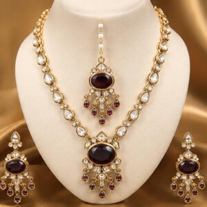 May include: A gold-toned jewelry set consisting of a necklace, a pair of earrings, and a maang tikka. Each piece features a large oval-shaped dark maroon stone surrounded by smaller clear stones, with dangling maroon teardrop accents throughout.