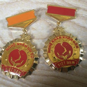 May include: Two gold-toned circular medals with scalloped edges, each featuring a red center with a map of Vietnam and hands. The left medal has an orange top bar and text reading "NGHỆ NHÂN ƯU TÚ VIỆT NAM". The right has a red top bar and "NGHỆ NHÂN NHÂN DÂN VIỆT NAM".