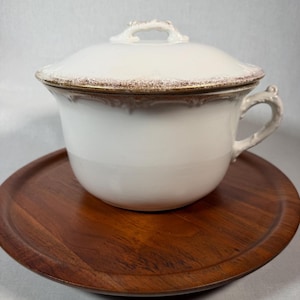 May include: A white ceramic chamber pot with a matching lid, featuring a gold-toned speckled rim and a decorative handle. The vessel has a smooth, rounded body and a slightly flared top edge, showcasing a classic design with subtle embossed detailing.