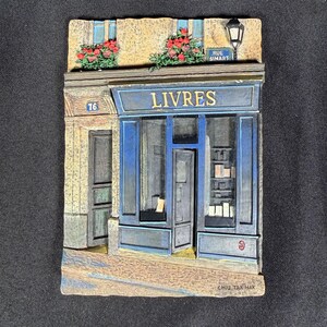 May include: A three-dimensional wall plaque depicting a bookstore storefront with a blue facade, the word LIVRES in gold lettering, a street sign reading RUE SIMART, red flower boxes, a black lantern, and the number 76 on a beige stone building.