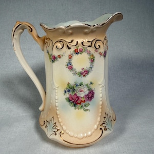 May include: A porcelain pitcher with a cream-colored base, featuring a scalloped rim and a curved handle. The body is decorated with hand-painted floral wreaths and bouquets in pink, purple, and green, accented with gold trim and beaded detailing.