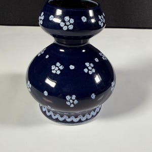 May include: A dark blue ceramic vase with a bulbous body and a flared neck. The surface is decorated with small, light blue painted flower clusters and a scalloped light blue pattern around the base. The finish is smooth and glossy.