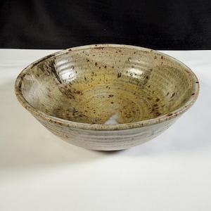 May include: A round ceramic bowl with a wide rim and a tapered base. The interior features a mottled glaze in shades of tan, beige, and brown with dark speckles. The exterior has a textured, ribbed surface with a light cream and brown finish.