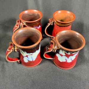 May include: A set of four ceramic mugs featuring a glossy red glaze with dark brown rims and a distinctive white drip pattern near the base. Each mug has a curved handle with a decorative, textured detail at the top, showcasing a handcrafted finish.