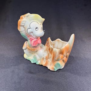 May include: A ceramic figurine planter featuring a cartoon character wearing a hat and a red neckerchief, standing next to a hollow, tree-stump-shaped vessel. The piece is finished in a glossy glaze with pastel orange, green, and yellow accents.