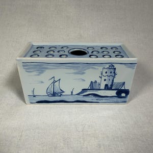 May include: A rectangular ceramic flower frog vase featuring a white base with blue hand-painted illustrations of a sailboat and a coastal tower. The top surface contains multiple circular openings for arranging stems, including one larger central hole.