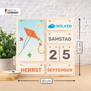 Könnte beinhalten: Ein quadratischer hölzerner immerwährender Kalender mit den Maßen 30 cm mal 30 cm. Er zeigt ein hellblaues Feld mit einer roten Drachenillustration sowie kleinere Felder mit dem Text WOLKEN, SAMSTAG, 25, HERBST und SEPTEMBER.