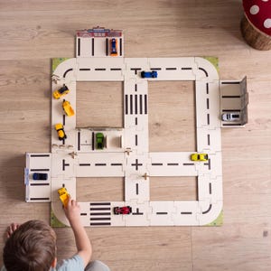 Könnte beinhalten: Ein modulares Spielzeugstraßensystem aus hellen Puzzleteilen mit schwarzen Straßenmarkierungen, einschließlich Kreuzungen, Zebrastreifen und Parkplätzen. Mehrere kleine Spielzeugautos in Gelb, Blau, Grün, Rot und Weiß stehen auf den Bahnen.
