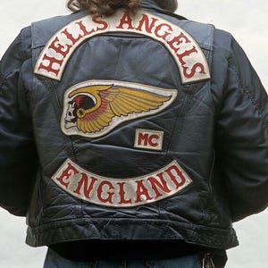 May include: A black leather vest featuring embroidered patches on the back. The top patch reads HELLS ANGELS in red text. A central patch displays a winged skull logo. A smaller patch reads MC, and a bottom curved patch reads ENGLAND in red text.