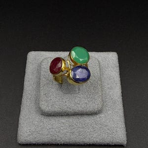 Puede incluir: Un anillo de tono dorado con tres piedras preciosas redondas y facetadas en rojo, verde y azul. Las piedras están dispuestas en un grupo sobre una banda de metal sencilla, mostrando un acabado pulido y un contraste de color definido.