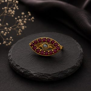 May include: A gold ring shaped like an eye, featuring a central round grey stone surrounded by a halo of small, deep pink circular gemstones. The ring has a pointed, almond-like silhouette and is crafted with a polished metallic finish.