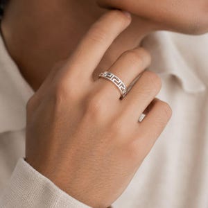 Könnte beinhalten: Ein silberner Ring mit einem durchgehenden, ausgeschnittenen griechischen Mäandermuster um das Band. Das Metall hat eine polierte Oberfläche und das Design besteht aus sich wiederholenden geometrischen Quadratmotiven, die einen Durchbruch-Effekt erzeugen.
