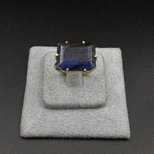 Puede incluir: Un anillo de oro con una gran piedra preciosa rectangular de color azul oscuro facetada. La piedra está montada en un sencillo engaste de garras de oro, con cuatro garras visibles que aseguran las esquinas de la piedra azul profunda.
