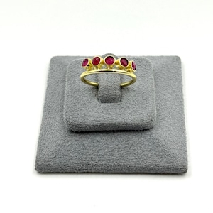 May include: A gold ring featuring five small, round, deep pinkish-red cabochon stones set in a row along the top of the band. The stones are held in place by individual gold bezels, creating a uniform, linear arrangement across the finger band.
