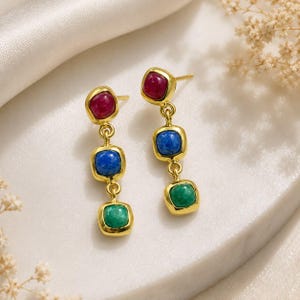 May include: A pair of gold-tone drop earrings featuring three stacked, square-shaped cabochon stones in descending order of colour: a deep red top stone, a vibrant blue middle stone, and a bright green bottom stone, all set in polished metal frames.