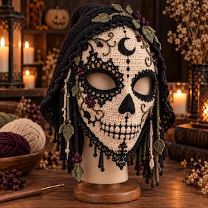 Puede incluir: Una máscara de calavera de ganchillo con cara beige, cuencas oculares, nariz y dientes bordados en negro. La máscara está adornada con una capucha negra, hojas de ganchillo verdes y pequeñas bayas de color rojo oscuro, con una luna creciente negra en la frente.