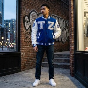 Könnte beinhalten: Eine blau-weiße Collegejacke mit den Buchstaben T und Z auf der Brust, der Zahl 3 auf dem rechten Ärmel, der Zahl 5 auf dem linken Ärmel und dem Text COB '03 auf der Vorderseite, kombiniert mit einer dunkelblauen Jeans und weißen Sneakern.