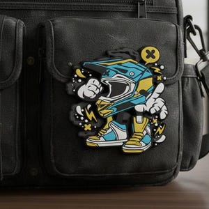 May include: A black messenger bag featuring a rubberized patch of a cartoon character wearing a blue and yellow motocross helmet and matching high-top sneakers. The character has white gloved hands, one pointing upward, with a yellow speech bubble containing an X.