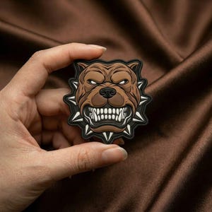 Pode incluir: Um emblema de borracha castanho e preto com uma ilustração estilizada de uma cabeça de cão a rosnar com os dentes à mostra, rodeado por uma série de espigões afiados dispostos num padrão circular à volta da cara do cão.