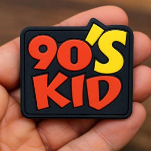 May include: A rectangular black patch featuring the text 90'S KID in raised lettering. The numbers 90 and the word KID are in a bold orange color, while the apostrophe and the letter S are in a bright yellow color against the black background.