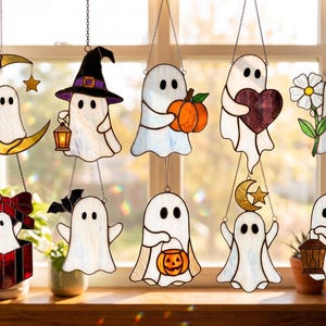 May include: A collection of ten stained glass ghost ornaments hanging in a window. Each ghost features white glass with black eyes, depicted with various accessories including a yellow crescent moon, witch hat, pumpkin, heart, flower, and lantern.