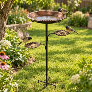 May include: A black metal bird feeder and bath station featuring a top copper-coloured basin for water and two lower scalloped trays for birdseed. The structure stands on a four-legged base, providing multiple levels for birds to land and feed.