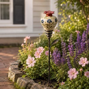 May include: A garden stake featuring a cream-colored ceramic vessel with blue floral patterns and green stems, topped with a red sculpted flower. The vessel sits atop a slender black metal rod with a single metal leaf attached to the stem.