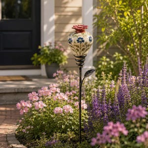 May include: A garden stake featuring a cream-colored ceramic vessel with blue floral patterns and green stems, topped with a red flower-shaped finial. The vessel sits on a black metal rod with a single metal leaf attached to the stem.