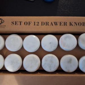 May include: A set of 12 round drawer knobs made of white marble with subtle grey veining, arranged in two rows of six inside a brown cardboard box. The box lid features the text "CASA DECOR" and "SET OF 12 DRAWER KNOBS" printed in black.