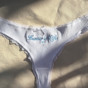 May include: White ribbed thong underwear featuring delicate lace trim along the edges and a small white bow at the center waist. The front is embroidered with the blue text Hunter's Wife above a small blue heart icon, creating a detailed design.