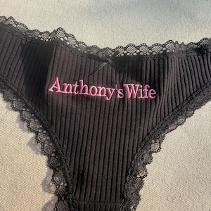 May include: Black ribbed underwear featuring black lace trim along the edges and a small black bow at the center waist. The text "Anthony's Wife" is embroidered in pink thread across the front center of the garment.
