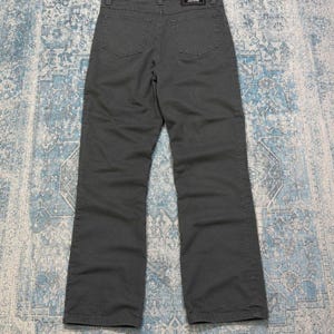 May include: A pair of charcoal gray denim jeans featuring a straight-leg cut, two back pockets, and a black rectangular leather patch on the waistband with the white text Versace Jeans Couture and a central Medusa head logo emblem.
