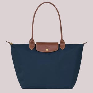 May include: A navy blue tote bag featuring a brown leather flap closure with a gold-tone snap button. The bag has two long, thin brown leather handles attached to the top. The body of the bag is made of a smooth, solid-colored fabric material.