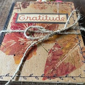 May include: A rectangular journal with a tan cover featuring printed red and orange autumn leaf patterns, black speckles, and decorative black stitching along the edges. The word Gratitude is printed on a label, and the book is tied with twine.
