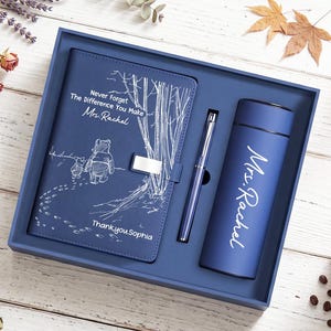 Op de afbeelding: Een blauwe geschenkset met een notitieboek, een pen en een beker. Het notitieboek heeft een witte lijntekening van Winnie de Poeh en Knorretje, met de tekst "Never Forget The Difference You Make Mrs. Rachel" en "Thank you Sophia". De beker is bedrukt met "Mrs. Rachel".