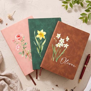 Peut inclure: Trois carnets rectangulaires aux couvertures lisses rose, vert foncé et marron. Le carnet rose présente une illustration de rose, le vert une jonquille jaune et le marron des fleurs de narcisse blanches avec le prénom Oliver inscrit dessus.