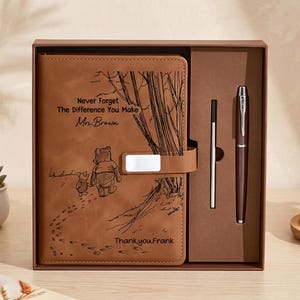 K&ouml;nnte beinhalten: Ein beigefarbenes Notizbuch in Lederoptik mit Magnetverschluss, graviert mit einer Illustration von Winnie Puuh und Ferkel, dem Text "Never Forget The Difference You Make", "Mrs. Brown" und "Thank you, Frank", sowie einem passenden Stift in einer Box.