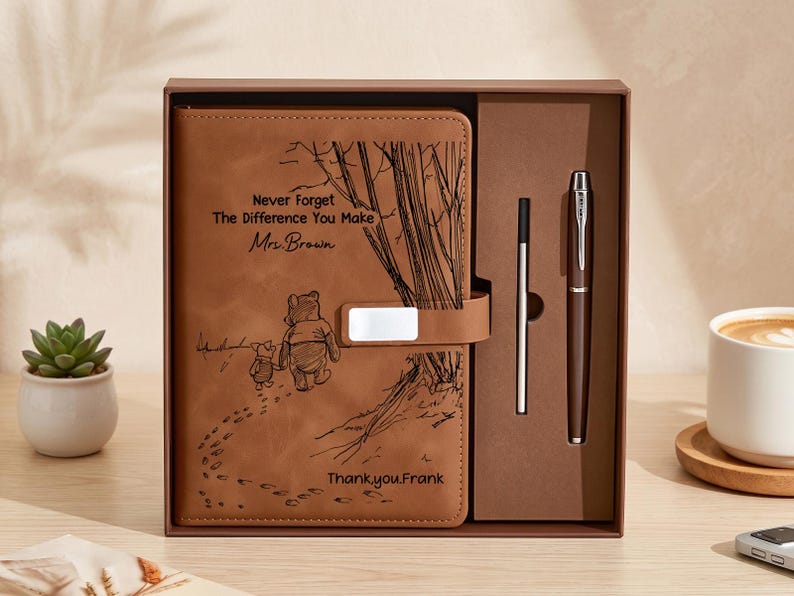 K&ouml;nnte beinhalten: Ein beigefarbenes Notizbuch in Lederoptik mit Magnetverschluss, graviert mit einer Illustration von Winnie Puuh und Ferkel, dem Text "Never Forget The Difference You Make", "Mrs. Brown" und "Thank you, Frank", sowie einem passenden Stift in einer Box.