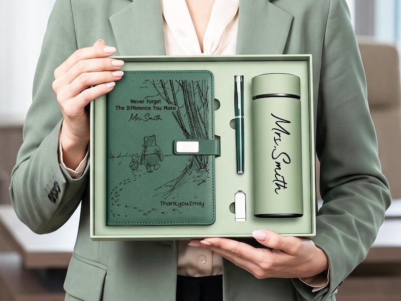 K&ouml;nnte beinhalten: Ein salbeigr&uuml;nes Geschenkset bestehend aus einem Notizbuch, einem Stift, einem USB-Stick und einem Becher. Das Notizbuch zeigt eine Winnie Puuh-Illustration mit dem Text "Never Forget The Difference You Make, Mrs. Smith, Thank you Emily." Der Becher ist mit "Mrs. Smith" bedruckt.