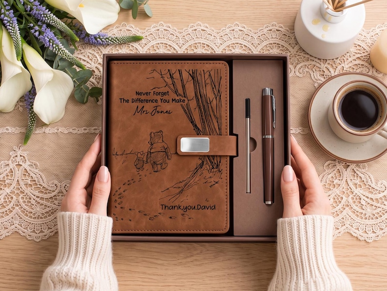 K&ouml;nnte beinhalten: Ein braunes Notizbuch in Lederoptik und ein Stift in einer Geschenkbox. Der Einband zeigt eine Lasergravur von Winnie Puuh und Ferkel sowie den Text "Never Forget The Difference You Make", "Mrs. Jones" und "Thank you David".